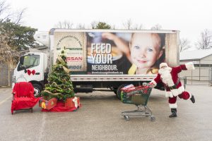 Santa Claus with a shopping cart full of donations next to a Christmas tree and in front of The Mississauga Food Bank truck.